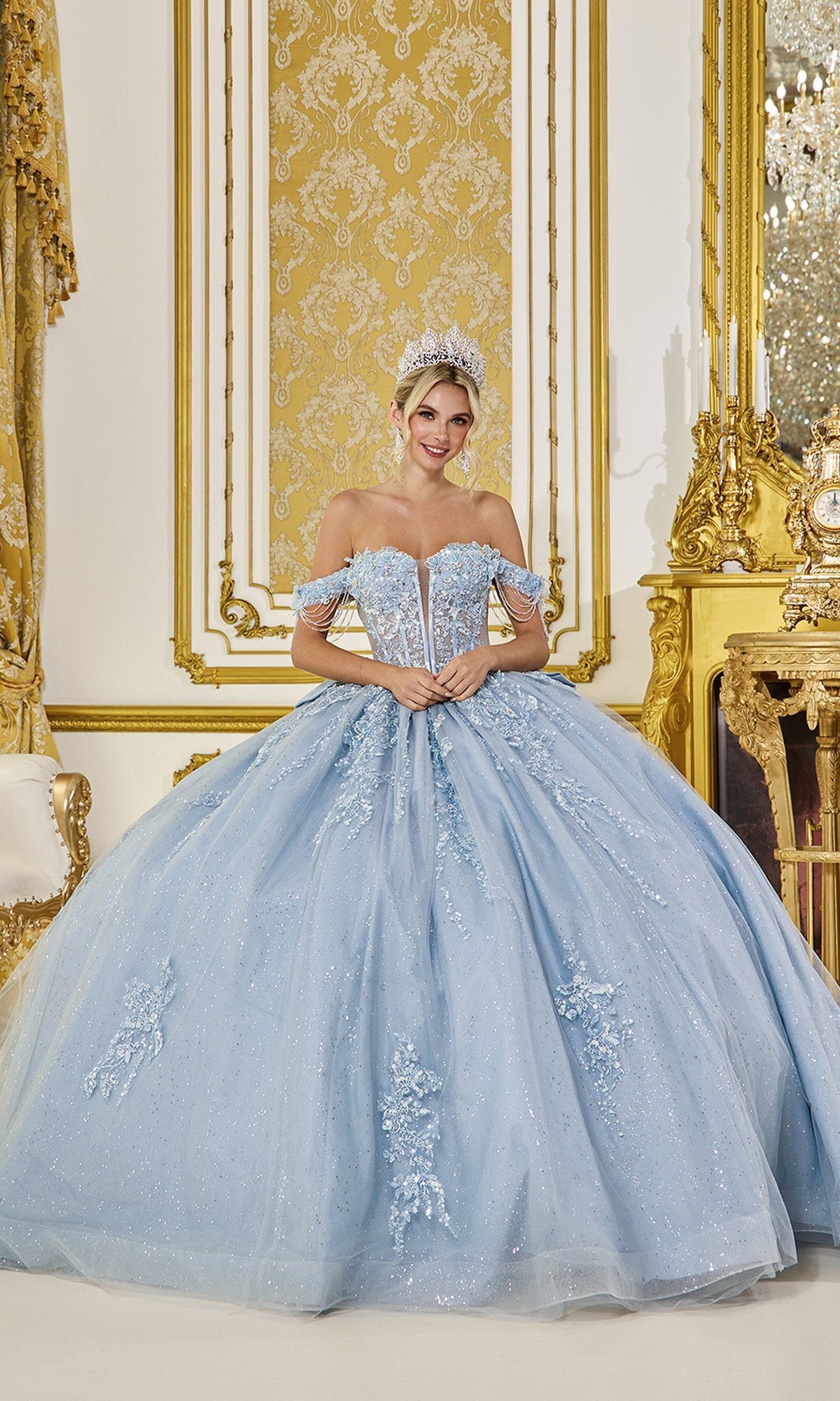 A woman wearing the Dusty Blue Quinceanera Dress 5092 By Dancing Queen and a tiara stands in an ornate gold and white room.